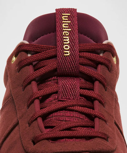 Maroon sneaker with gold laces and brand logo on a gray background
