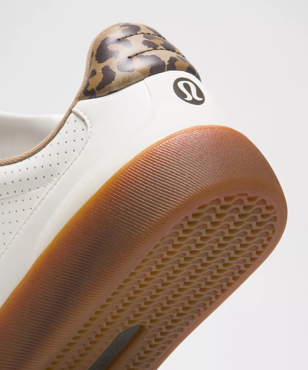 Close-up of a white sneaker with a brown sole and leopard print heel tab on a light gray background.