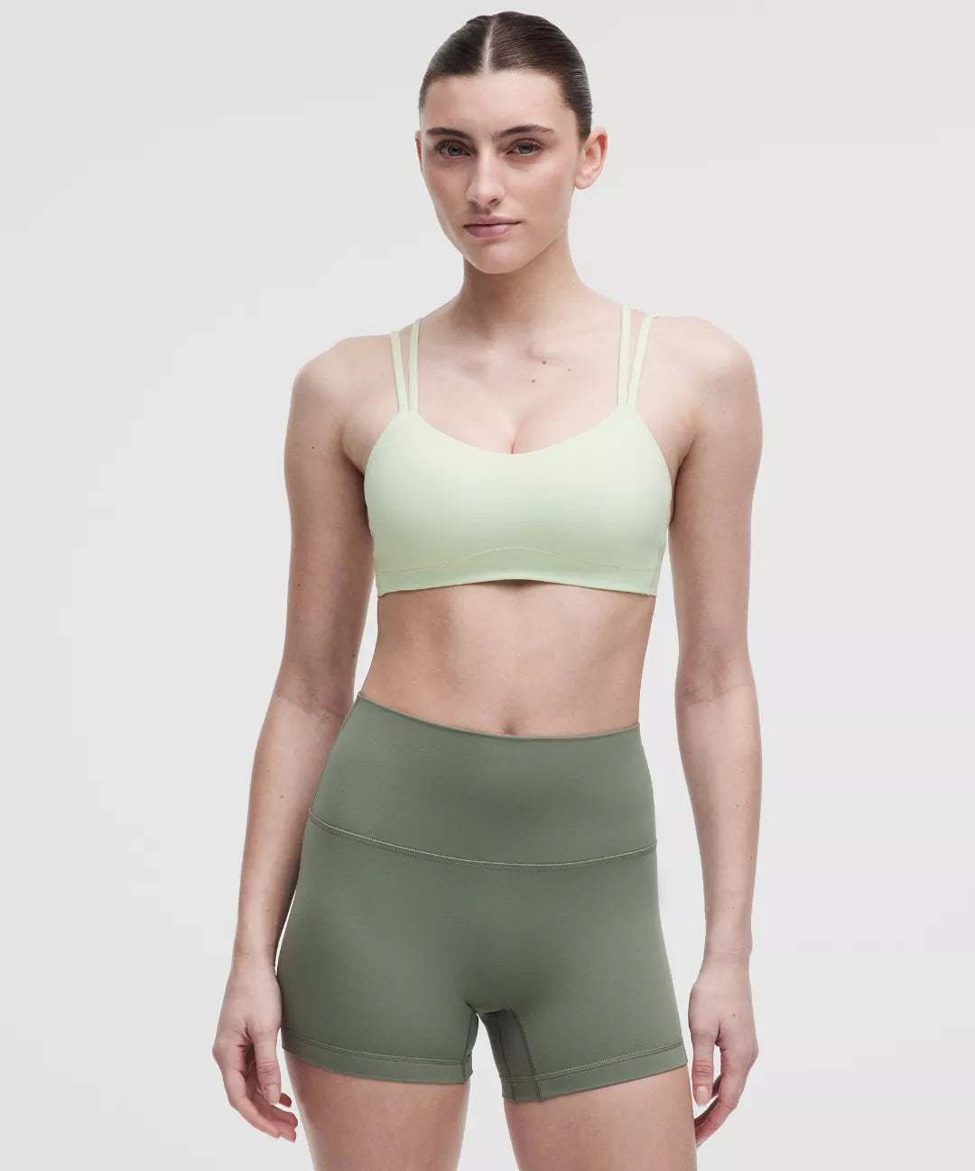 Woman wearing a light green sports bra and olive green shorts on a white background