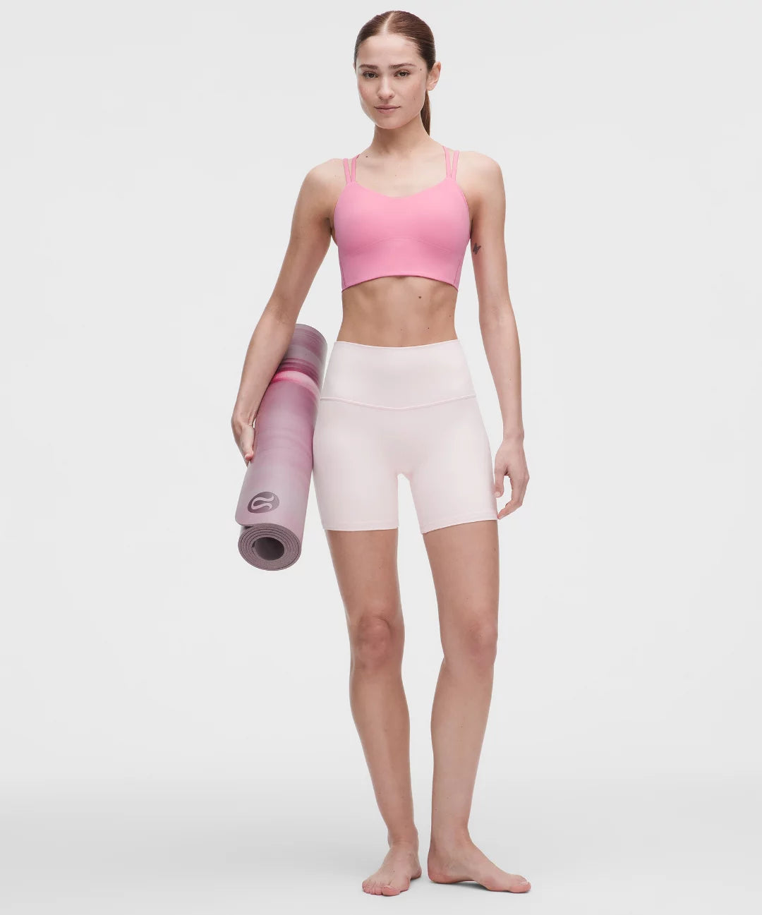 Woman holding a pink yoga mat wearing a pink sports bra and white shorts on a light gray background