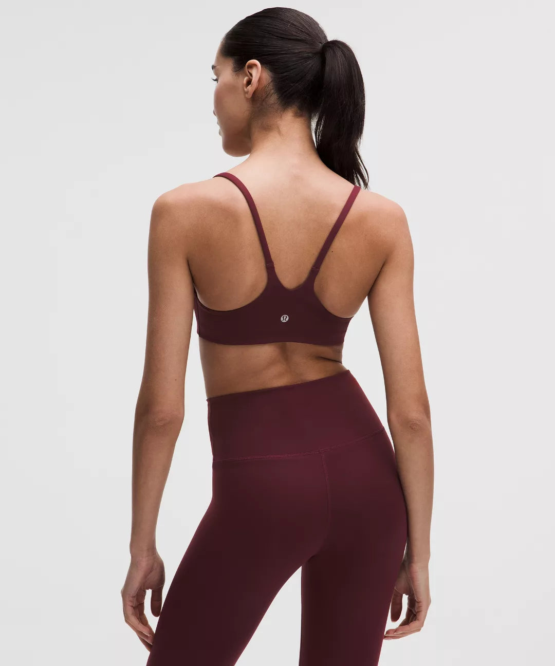 Woman wearing a maroon sports bra and leggings at back side on a light gray background 
