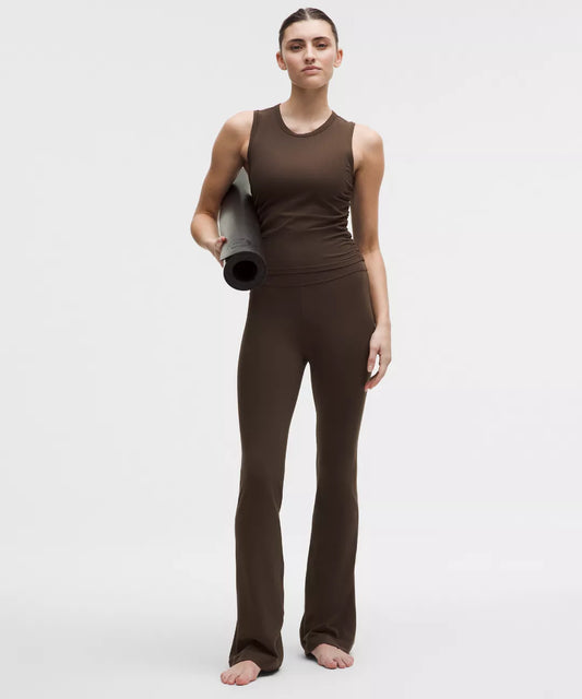 Person wearing a brown sleeveless top and pants holding a yoga mat on a white background
