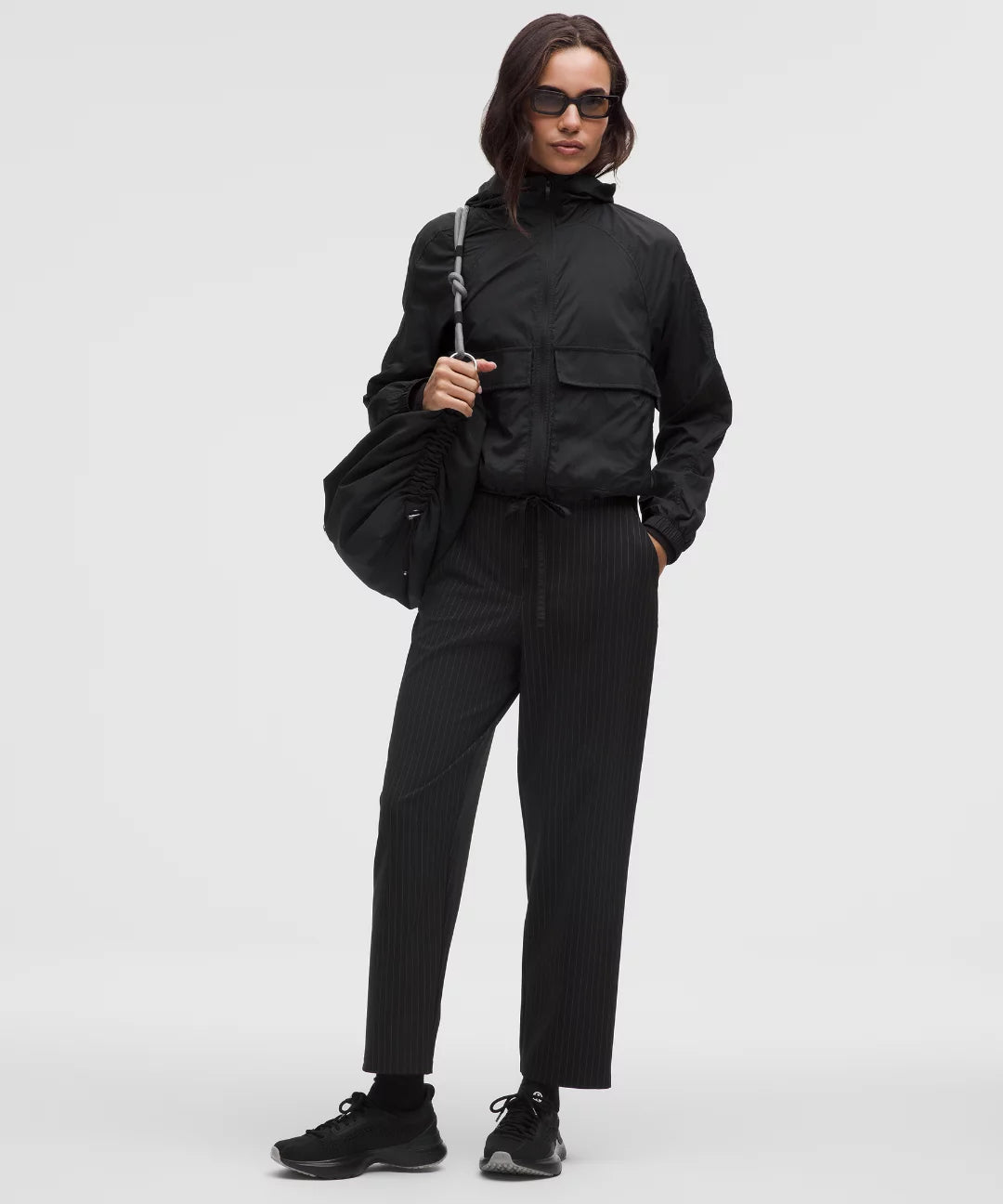 Person wearing a black outfit with sunglasses holding a black bag on a white background