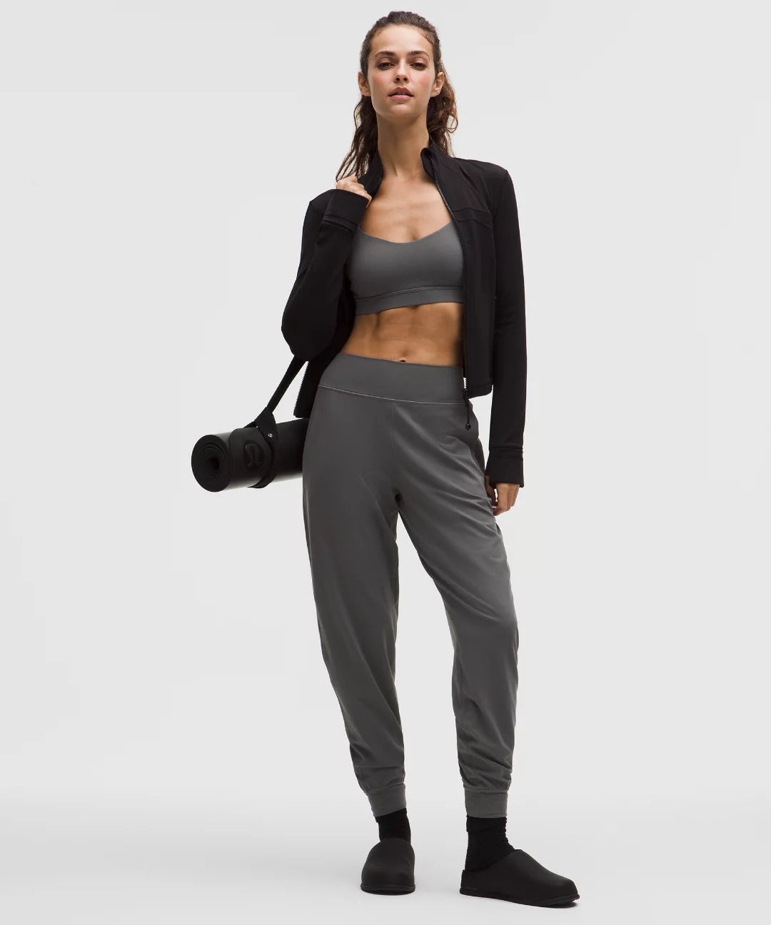 Woman in gray athletic wear holding a black yoga mat on a white background