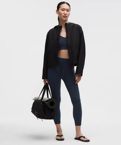 Woman holding a black duffel bag wearing a black jacket, blue leggings, and black sandals on a white background