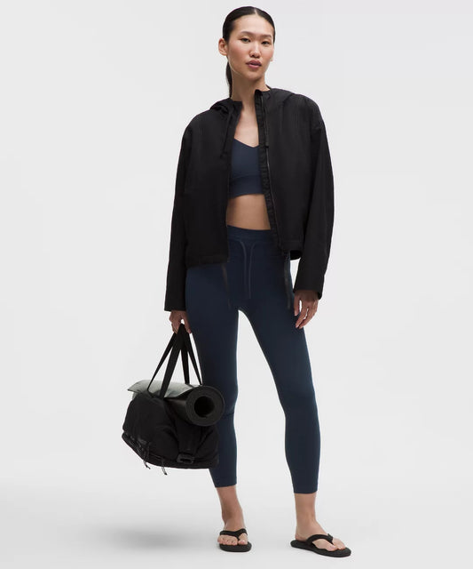 Woman holding a black duffel bag wearing a black jacket, blue leggings, and black sandals on a white background