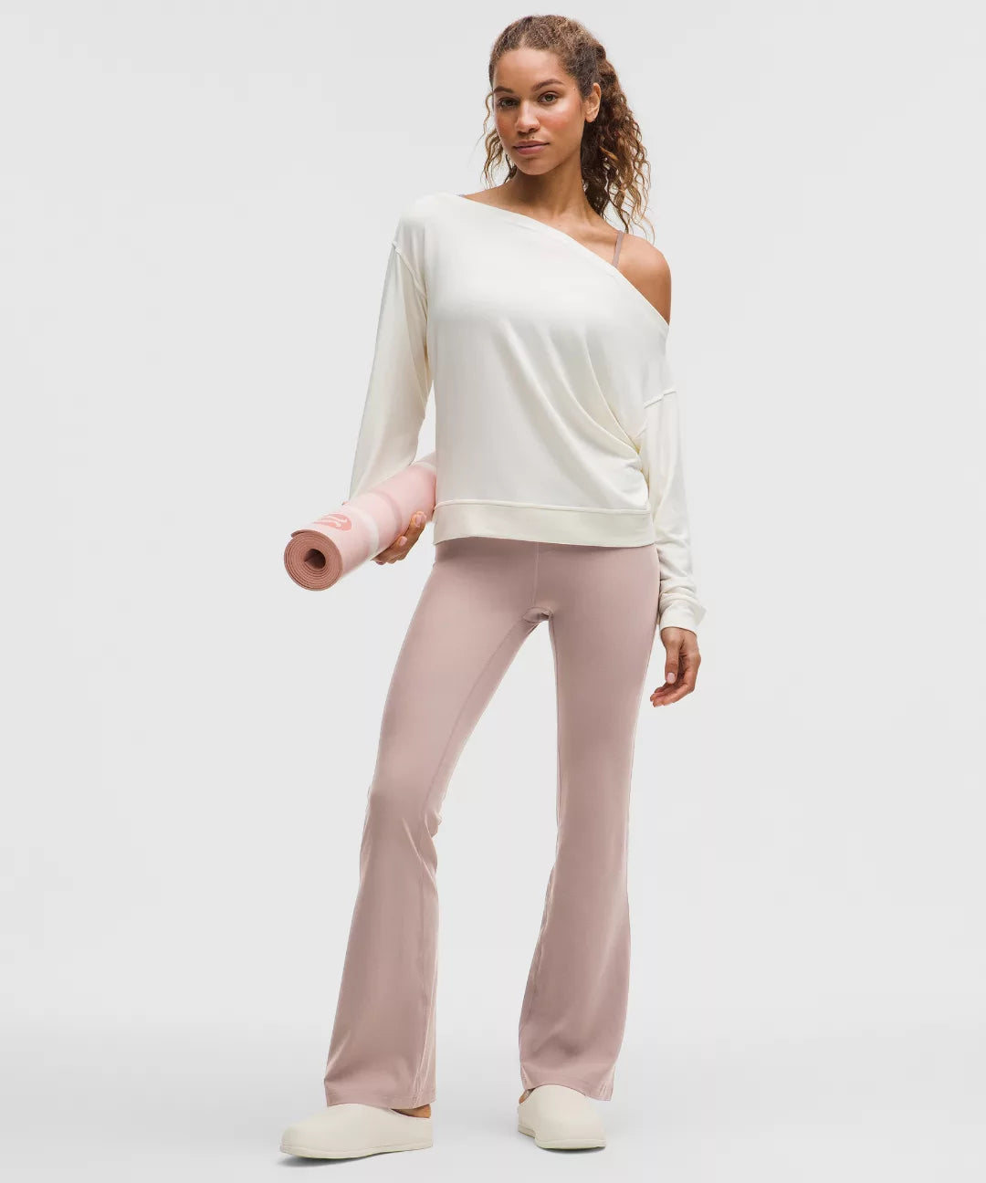 Woman holding a pink yoga mat wearing a white top and pink pants on a light gray background