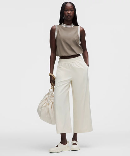 Woman wearing a beige sleeveless top and white pants, holding a beige handbag on a white background