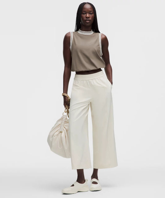 Woman wearing a beige sleeveless top and white pants, holding a beige handbag on a white background
