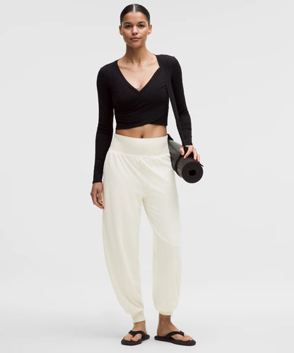 Woman holding a yoga mat wearing a black long-sleeve top and white pants on a plain background