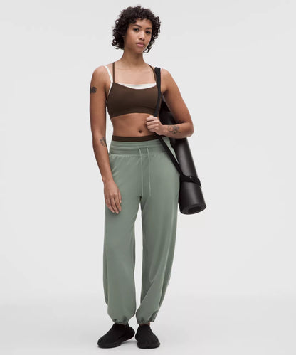 Woman holding a yoga mat wearing a brown sports bra and green pants on a white background