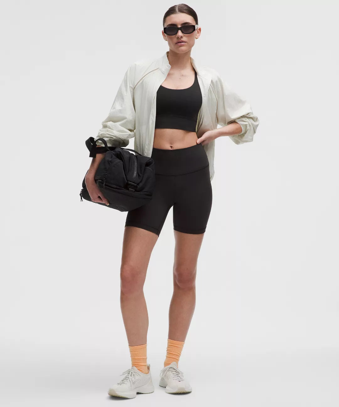 Person wearing a black athletic outfit with a white jacket and sunglasses, holding a black bag on a light gray background.