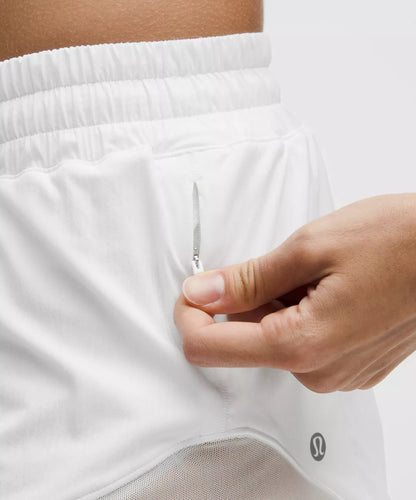 Close-up of a person wearing white shorts with a hand zipping them up, on a light gray background.