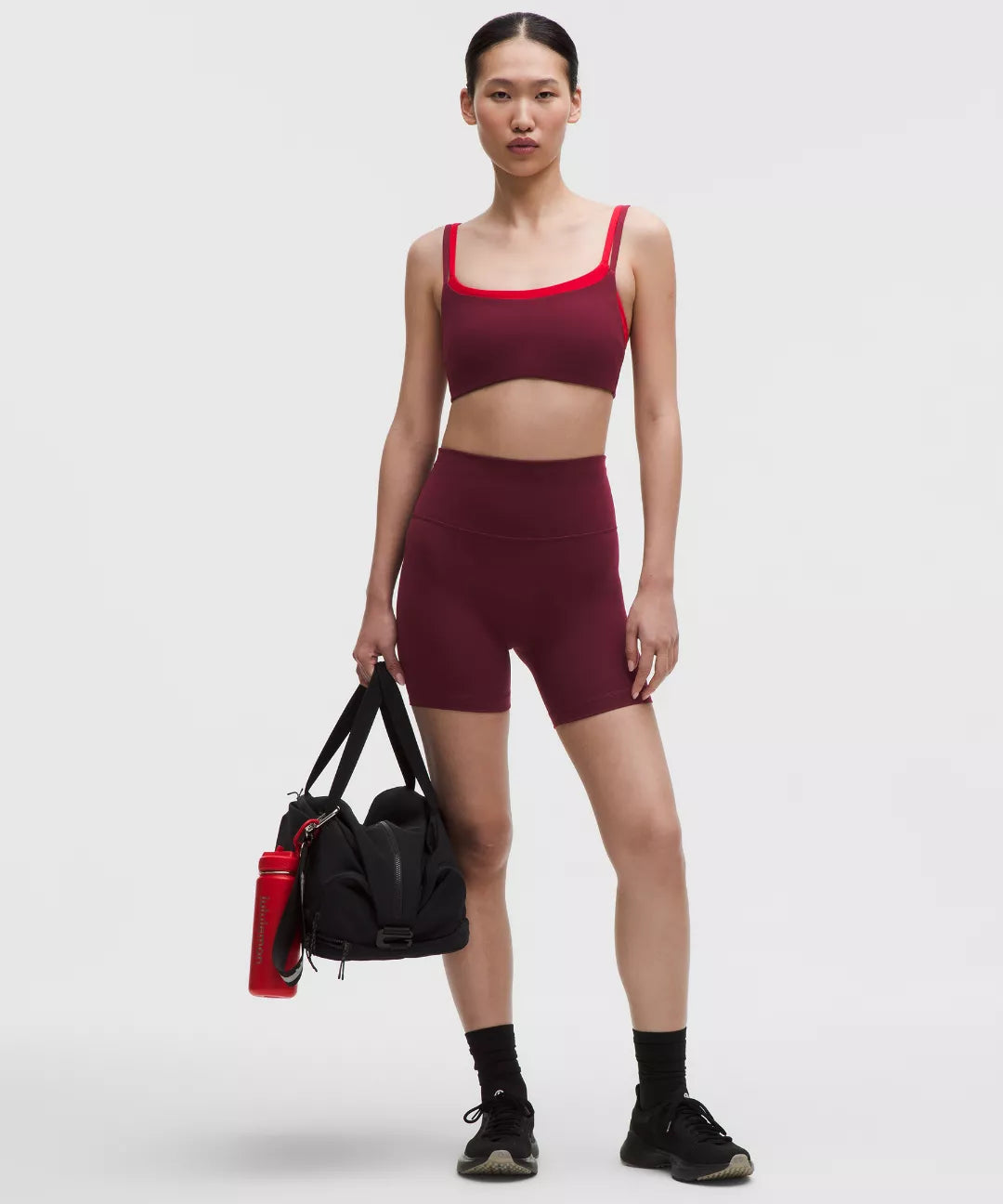 Woman in maroon athletic outfit holding a black bag and red water bottle on a white background